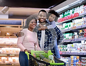 Portrait of positive black family with shopping cart smiling and looking at camera at modern supermarket