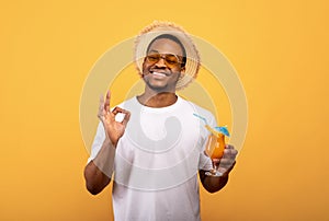 Portrait of positive black man drinking refreshing cocktail, having beach party, showing okay gesture, yellow background