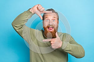 Portrait of positive carefree funny man with red hairstyle khaki pullover fingers showing camera cadre isolated on blue