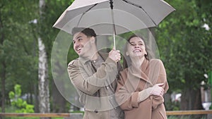 Portrait of positive Caucasian handsome man smiling and hugging loving girlfriend on rainy day outdoors. Middle shot of