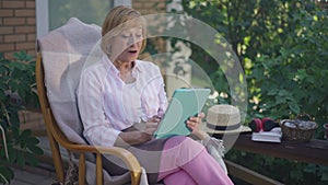 Portrait of positive Caucasian senior woman scrolling social media surfing Internet on tablet sitting on porch outdoors