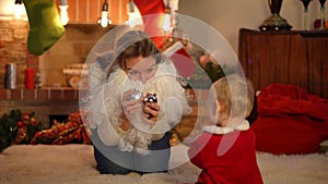 Portrait of positive Caucasian woman playing with Christmas balls taking off Santa beard talking to baby girl sitting on