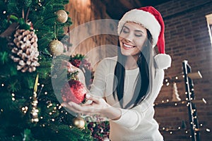 Portrait of positive cheerful girl in santa claus headwear hang christmas ball on evergreen tree for x-mas newyear