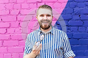 Portrait of positive cheerful young stylish man with mustache and beard and glasses in hands posing against blue-violet