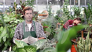 Portrait of positive confident adult floriculturist working in gardening store, talking among greenery of ornamental