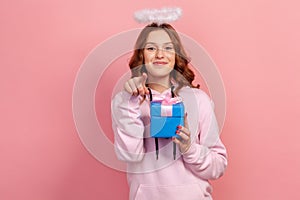 Portrait of positive curly haired teenage girl in hoodie with halo on head holding wrapped gift box and pointing finger on camera