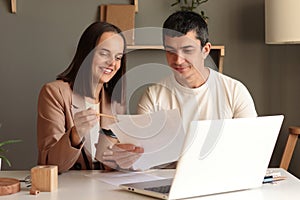 Portrait of positive delighted man and woman sitting at table together in front of laptop, working together with papers, studying