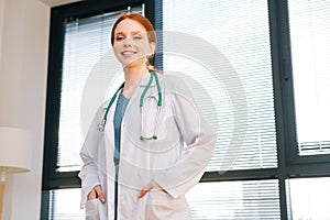 Portrait of positive female doctor in white coat standing on background of window in sunny day in light medical clinic