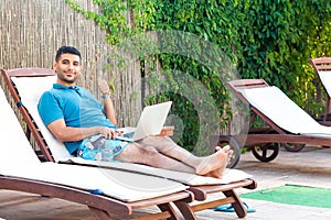 Portrait of positive handsome bearded young adult freelancer man in blue t-shirt and shorts lying on cozy sunbed with laptop on photo