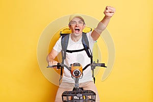 Portrait of positive happy optimistic deliveryman on bicycle wearing white T-shirt and cap isolated over yellow background, raised