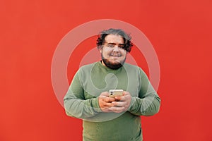 Portrait of a positive hispanic man with curly hair, smiling and holding a mobile phone. Cheerful young obese guy is using his