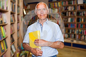 Portrait of positive intelligent older man in library with book in hands