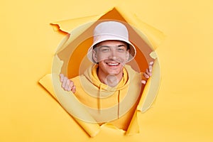 Portrait of positive joyful man wearing casual hoodie and panama looking through torn hole in yellow paper, looking at camera with