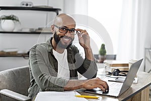Portrait of positive latino male freelancer working on laptop and smiling at camera, sitting at desk at home, free space
