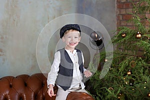 Portrait of positive little boy in cap against backdrop of Christmas tree with garland lights. Sincere child emotions. Closeup fac