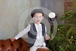 Portrait of positive little boy in cap against backdrop of Christmas tree with garland lights. Sincere child emotions. Closeup fac