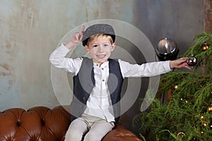 Portrait of positive little boy in cap against backdrop of Christmas tree with garland lights. Sincere child emotions. Closeup fac