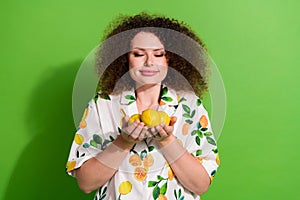 Portrait of positive lovely girl closed eyes arms hold smell fresh juicy lemons  on green color background