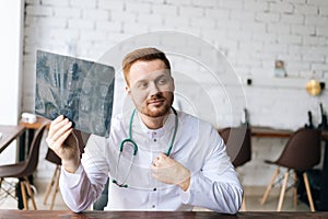 Portrait of positive male doctor in white uniform examining brain computerized tomography scan sitting at desk in