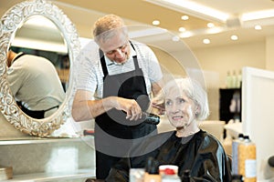 Portrait of positive man hairdresser cutting elderly woman hair in salon