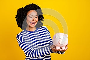 Portrait of positive nice girl with afro hairstyle wear striped pullover look at money box in hands isolated on yellow