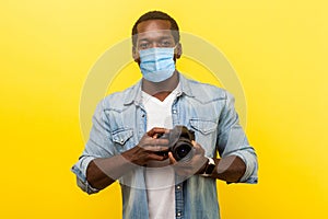 Portrait of positive photographer, man with medical mask holding professional digital dslr camera and looking with toothy smile,