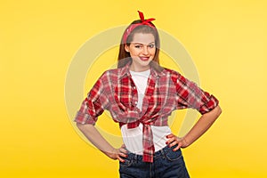 Portrait of positive pinup girl with bright makeup, red lipstick, in checkered shirt and headband, holding hands on hips