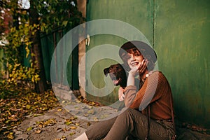 Portrait of positive rural girl in vintage clothes and hat sitting with dog on the ground at country house in autumn beautiful day