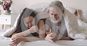 Portrait of positive senior Caucasian couple lying in bed under blanket, chatting and laughing. Smiling mature man and