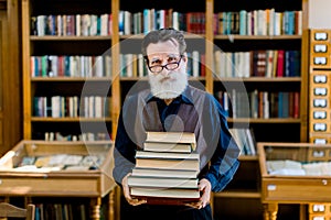 Portrait of positive smart old bearded man in dark shirt and leather vest, library worker, teacher, working in library