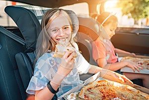 Portrait of positive smiling girl eating just cooked italian pizza sitting with a sister on car back seat in child car seats. photo