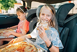 Portrait of positive smiling girl eating just cooked italian pizza sitting with a sister on car back seat in child car seats. photo