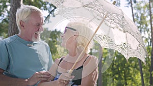 Portrait of positive smiling mature woman in sunglasses standing outdoors under the white parasol. Senior man meeting