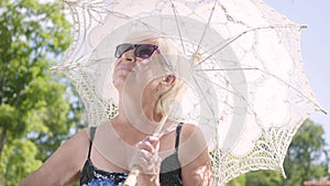 Portrait of positive smiling mature woman in sunglasses standing in the park under the white parasol looking around and