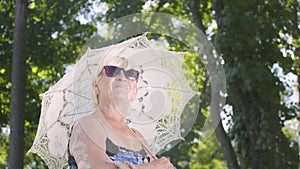 Portrait of positive smiling mature woman in sunglasses standing in the park under the white umbrella looking around