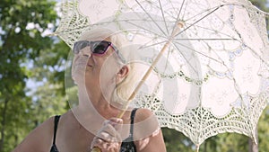 Portrait of positive smiling mature woman in sunglasses standing in the park under the white umbrella looking around