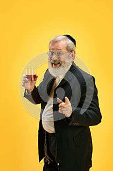 Portrait of positive, smiling senior man holds glass of whiskey and pointing to camera against yellow background. Happy