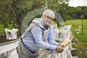 Portrait of a positive smiling senior man walking in a summer park with a book in his hands.