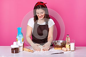 Portrait of positive woman dresses white t shirt and apron, has dirty face with flour, kneads dough, loooks down and smiles,