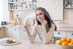 Portrait of positive woman drinking water from glass, sitting at table in kitchen interior and smiling at camera