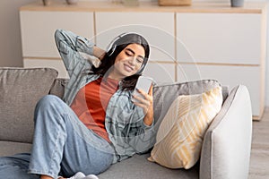 Portrait of positive young Arab woman in headphones chilling on sofa, listening to music on mobile device at home