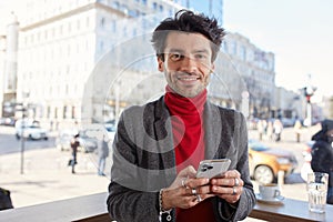 Portrait of positive young attractive brown haired male with trendy haircut smiling cheerfully at camera and keeping smartphone in