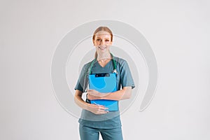 Portrait of positive young female physician in medical uniform holding clipboard and looking at camera.