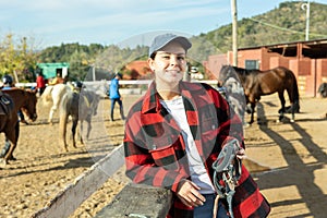 Portrait of positive young female stable worker holding horse halter or harness near wooden fence on ranch
