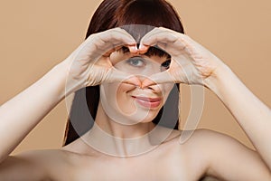 Portrait of positive young lady with healthy skin holding hands in the shape of heart near eye