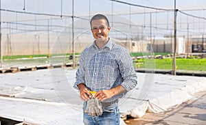 Portrait of positive young male worker posing at greenhouse on background with plantation of sprouts