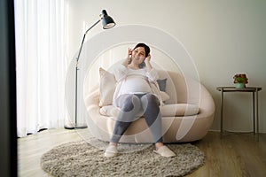 Portrait of pregnant Happy Asian woman sitting on sofa at home and using smart phone for listening music in headphones