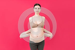 Portrait of pregnant woman putting on supporting bandage for reducing backache at pink background with copy space. Orthopedic