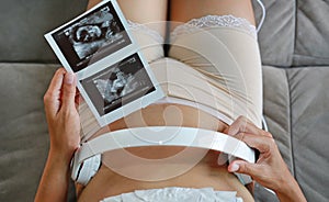 Portrait of pregnant woman sitting on sofa at home holding ultrasound film and listening music in headphones