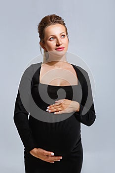 Portrait of a pregnant woman in a tight black dress on a gray background. The woman gently holds her stomach with her hands